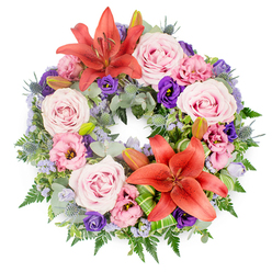 This overhead view captures a lush, circular sympathy wreath resting against a white background, its colours and textures working together in quiet harmony. Two vivid orange-red Asiatic lilies anchor the arrangement, one positioned toward the upper arc of the circle and the other toward the lower section, their open petals curving back to display dark stamens and smooth throats that seem to glow with warmth. Between and around them, numerous softly coloured roses in blush and pale lavender-pink tones are packed closely, their spiralling petals slightly cupped and velvety, suggesting tenderness and affectionate remembrance. Threaded among the roses, lisianthus blooms in gentle pink and rich, regal purple add a ruffled, almost fluttering texture as their layered petals drift outwards. Tiny lavender-blue filler flowers, similar to statice or limonium, are scattered delicately through the design like a light veil, easing the transition between the warmer oranges and cooler purples. The entire circle is supported by a deep bed of mixed greenery: rounded, silver-green eucalyptus, darker polished leaves, and feathery fern fronds overlap to create a thick, natural framework. Standing out from this foliage are blue-grey eryngium thistles, their spiky, textural heads and slender stems adding a modern, sculptural note that still feels completely appropriate for a farewell tribute. Here and there, closed lily buds rest quietly among the leaves, a subtle reminder of life continuing. At the base of one lily, bright green leaves are artfully folded and wrapped, betraying the careful hand of a local Stanmore florist. It is the sort of wreath Flower Delivery Stanmore might prepare for a thoughtful condolence piece delivered to a family home near Stanmore Country Park or a chapel near Canons Park station.