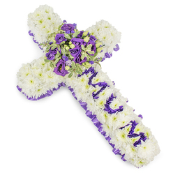 A deeply reverent memorial cross formed from tightly packed white chrysanthemums, every bloom positioned to create a plush, even surface that suggests both purity and solace. The cross is edged with a pleated purple satin ribbon whose ruffled texture introduces a refined, somber contrast to the snowy whites. Spelled down the vertical beam, the letters MUM appear in rich purple flower heads - likely small spray chrysanthemums or eustoma - their saturated colour declaring a personal, intimate tribute. The intersection of the cross is anchored by a generous central spray: deep violet roses with tight, velvety petals sit beside soft lavender lisianthus, their frilled edges and translucent layers catching the light. Variegated foliage with lighter margins and darker centres, perhaps pittosporum, gives depth and a cooling green tone, while tiny green berries add an understated, natural sparkle. The image is photographed on a clean white backdrop, allowing the purple and white palette to breathe and the arrangement's craftsmanship to read clearly. I picture this piece arriving at a service in Stanmore - close to Bentley Priory or the Tube - where the restrained scent of the blooms and the tactile fullness of the chrysanthemums provide comfort in quiet reflection. As a local artisan, Flower Delivery Stanmore often creates memorials like this for families seeking a dignified expression of grief and remembrance in the Stanmore and Harrow area.