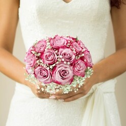 This hand-tied bouquet is a poetic highlight of a Stanmore bride's ensemble, capturing the soft interplay of colour and texture beneath the gentle glow of afternoon light. Nearly a dozen fresh roses unfurl in a captivating spectrum, blushing from creamy pink at their hearts to vivid magenta at the edges. Artfully interlaced swathes of baby's breath infuse the arrangement with ethereal whiteness, a misty cloud that dances around the more structured blooms-a familiar sight in Stanmore's picturesque ceremony venues. The bride's tanned arms and elegantly manicured nails offer a warm, steady cradle, set just above a softly tied sash detail that completes her sleeveless gown. Delicate lacework graces the bodice, providing a tactile counterpoint to the velvety roses. The background, comfortably blurred, suggests peace and allows full celebration of the bouquet's craftsmanship. This arrangement, emblematic of a Stanmore wedding, embodies cherished memories and the promise of new journeys-an artistic flourish that defines wedding-day beauty across the local community.