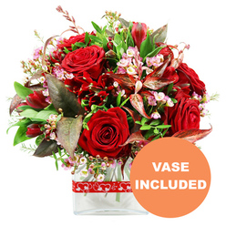 An elegant, passion-filled floral arrangement rests in a clear glass vase, set against a crisp white background that highlights every detail, much like a bouquet ready to go out from a Stanmore florist's workbench. The focal point is a cluster of velvety red roses, each bloom at a different stage, from tightly furled buds that feel full of anticipation to more open flowers revealing their layered hearts. Weaved amongst them are smaller, equally rich deep red flowers, reminiscent of alstroemeria or similar multi-petalled stems, building texture and giving the eye plenty to explore. Threaded delicately through the red tones are sprays of tiny, soft pink and creamy white waxflowers or limonium, their petite blossoms forming clouds of airy lightness, softening the intensity of the colour palette. A lush frame of greenery surrounds the blooms: wide, glossy green leaves, finer stems of lighter foliage, and distinctive burgundy and reddish-brown leaves with subtle veining that echo the warmth of the roses and add an extra layer of sophistication. The bouquet is shaped into a generous, rounded dome, suggesting a hand-tied style crafted with care and precision. It is anchored in a simple, modern rectangular glass vase, the stems arranged cleanly in the clear water, giving the design a contemporary, uncluttered feel. A vivid red ribbon, decorated with a repeating motif of white hearts, encircles the vase, reinforcing the bouquet's romantic intention-perfect for an anniversary dinner near Stanmore Hill or a surprise delivery to a home close to Stanmore Country Park. In the lower right corner of the image, a strong orange circular overlay bears the text VASE INCLUDED in neat white uppercase letters, signalling that the arrangement arrives complete and ready to display the moment it is received.