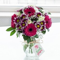 In this image, a lovingly crafted floral arrangement glows at the centre of a bright, softly blurred white backdrop, much like a windowsill catching late morning light in a quiet Stanmore home. The bouquet overflows from a clear, subtly textured glass vase, where you can see the neatly trimmed stems gathered together. Dominating the display are generous gerbera daisies in vivid shades of fuchsia and magenta, their dark, almost chocolate-coloured centres giving a striking focal point that feels both modern and exuberant. Surrounding them are intricate chrysanthemums with multi-layered petals, their outer edges painted in rich purple that gently melts into white, encircling sunny yellow hearts that recall carefree summer days in nearby Stanmore Country Park. Nestled among these are creamy white spray roses, some just beginning to unfurl, their soft, spiralled petals suggesting tenderness and new beginnings. Fine sprigs of white baby's breath thread between the larger blooms, lending an airy, sparkling quality, as if the bouquet is sprinkled with tiny stars. Deep green foliage provides contrast and structure, framing the brighter flowers and giving the arrangement a lush, garden-fresh feel, as though just cut from a well-tended back garden off Marsh Lane. The vase is finished with a rustic loop of natural twine tied in a simple bow, from which hangs a white gift tag delicately printed with a pink rose graphic and cursive lettering, hinting at a personal message without revealing the words. The overall mood is one of joyful celebration and heartfelt thoughtfulness, ideal for marking birthdays, anniversaries, or welcome-home moments with local flower delivery across Stanmore and the surrounding streets.