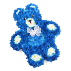 A three-dimensional floral teddy bear rests in the centre of a white background, its details standing out clearly thanks to the simple, uncluttered setting. The bear is constructed entirely from fresh flowers, with royal blue blooms serving as the main colour for the head, body, and limbs. These small, densely packed flowers, similar to miniature chrysanthemums, create a thick, soft texture that calls to mind the feel of a well-loved soft toy, the sort that might sit on a child's bed in a Stanmore family home. The blue petals are uniform and vivid, suggesting serenity and remembrance. Against this deep blue, the florist has used creamy white and faintly yellow-tinted blossoms to pick out the inner ears, rounded muzzle, and four oval paw pads, giving gentle definition and a playful, childlike charm. The teddy's eyes are made from dark, glossy discs that appear almost glassy, adding a lifelike sparkle, while a small black triangle forms the nose and a fine curved line below completes a peaceful, tender smile. Around the neck, a delicate bow made from sheer white mesh or organza is tied in a crisp, neat bow, its light, gauzy texture catching the light softly. The bear lies slightly angled, introducing a sense of movement and making the design feel more personal and approachable. This floral sculpture is clearly intended as a thoughtful tribute, particularly suited for remembering a baby boy or young child during funerals or memorial services in Stanmore. The balance of colours, the symmetry of the features, and the flawless shaping reveal a high level of craftsmanship, offering a touching symbol of love, innocence, and gentle farewell.