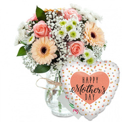 This photograph portrays a thoughtful Mother's Day gift set, styled as though ready to be delivered to a quiet home in Stanmore, perhaps just off Uxbridge Road or near the green spaces of Canons Park. The composition is clean and bright, with every element standing clearly against a white background. On the left rests a clear, straight-sided glass vase, its transparency revealing the gathered stems within. Around the neck, a length of light brown twine is tied in a simple bow, adding a touch of rustic personality that softens the formality of the glass. Rising from the vase is a lush, rounded bouquet of pastel flowers. Several peach-pink roses open in generous spirals, their petals layered like soft fabric. Large peach gerbera daisies punctuate the bouquet, their daisy-like petals framing bold, dark brown centres that provide a focal point. Intermingled among them are small white chrysanthemums with pale green cores and airy clusters of white baby's breath, which appear almost like fine, floating bubbles of light. Fresh green foliage threads through the blooms, framing the colours and suggesting the crisp scent of newly cut stems. Behind the bouquet, almost hidden, is a small white greeting card with hints of green and pink handwriting, bringing a personal whisper of Happy Mother's Day from child to mother. To the right of the vase a heart-shaped foil balloon stands proudly forward. Its glossy white surface is dotted with sparkling gold and silver spots, and in the middle a soft peach heart holds the words HAPPY Mother's DAY written in bold black script, the word Mother's drawn in graceful, calligraphic strokes. The overall impression is of joy, tenderness and gratitude, ideal for same-day Mother's Day flower delivery across Stanmore and neighbouring Harrow Weald.