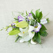 On a neutral, off-white fabric surface that looks like softly worn linen, a hand-tied bouquet lies gracefully on its side, its composition speaking of careful balance and quiet luxury. At the centre sit white Oriental lilies, some fully open with their broad, wax-like petals curling back from the centre, others held in sleek green buds that curve along the length of the arrangement. In the open lilies, the stamens stand out in warm yellow and orange tones, dusted with dark pollen, while the inner petals show subtle freckles that add personality to each bloom. Around these lilies weave lisianthus flowers in white and shades of purple, their ruffled petals edged in lilac and velvety violet. Some eustoma blossoms are just beginning to unfurl, while pointed buds tipped with purple peek from between the leaves, lending a sense of youth and unfolding romance. Tiny stars of white baby's breath are scattered throughout the bouquet, creating a soft, cloud-like veil that lightens the overall feel. Encasing the flowers is an assortment of greenery: glossy deep green leaves, airy fern-like fronds, and thin, linear blades that introduce movement and structure. The stems are gathered tightly at the base and wrapped with smooth white satin, embellished with minute pearl-like details that offer a refined sparkle. Created for a Stanmore bride or a couple celebrating an anniversary near the quiet streets off The Broadway, this bouquet feels intimate yet sophisticated, as if waiting on a polished table before being carried down the aisle, its appearance evoking purity, devotion, and gentle optimism for the shared life ahead.