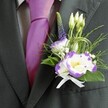 A close-up image shows the upper torso of a man dressed for a wedding or formal celebration in Stanmore, with the camera lingering on the rich detail of his attire and floral buttonhole. He wears a perfectly tailored dark charcoal grey suit jacket, its lapel clean and sharp against a softly patterned light grey check dress shirt. At his throat, a lustrous magenta silk tie is knotted with care, the sheen of the fabric catching the light and adding a bold streak of colour that feels both confident and joyful. Pinned to the left lapel is a carefully crafted boutonniere: a white lisianthus bloom edged in deep purple, its ruffled petals opening gently around a sunny yellow centre, supported by a second lisianthus head that echoes the same colour play. Around them cluster slender, closed white buds that speak of new beginnings, while a single upright stem of dark indigo veronica adds height and a subtle, almost wild-garden touch, like something gathered near Bentley Priory Nature Reserve. Tiny white filler flowers, reminiscent of baby's breath, nestle between fine feathery greenery and broader emerald leaves, creating a soft halo of texture. Loops of thin decorative green grass arc gracefully, giving the design a modern, almost sculpted feel, yet it remains romantic and understated. At the base, a neat white satin ribbon is tied in a tiny bow, securing the stems with a traditional flourish. The lighting is gentle, perhaps indoor afternoon light in a Stanmore venue, giving the scene a sense of quiet anticipation, as if the groom is moments away from walking down the aisle, carrying the fragrance and meaning of his flowers close to his heart.