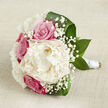 This hand-tied bouquet is displayed horizontally on a neutral, lightly textured fabric in soft beige, echoing the calm tones of a Stanmore home or boutique wedding venue. At its centre sits a lush creamy white peony, fully open and richly layered, each ruffled petal curling softly outward to form a voluminous, cloud-like bloom that immediately becomes the focal point. Circling this central flower are vibrant fuchsia-pink roses, their petals wound tightly in graceful spirals, the colour deep and passionate, bringing a sense of devotion that would feel at home in a ceremony near Stanmore Hill or along Uxbridge Road. Interlaced throughout the bouquet are fine sprays of gypsophila, or baby's breath, their tiny white blossoms creating a light, almost shimmering texture that contrasts with the bold, smooth rose petals and the soft fluffiness of the peony. Framing the bouquet, broad, dark green leaves arc gracefully around the blooms, their surfaces smooth and lightly reflective, giving the design structure and a fresh, garden-like feel, as though gathered from well-tended beds in a local Stanmore garden. The stems are gathered neatly and wrapped in a band of white satin ribbon, forming a sleek, polished handle that looks comfortable to hold for bridal portraits or aisle walks. Gentle, diffused lighting highlights the intricate details of each flower, accentuating the creamy tones of the peony and the vivid pink of the roses, while soft shadows add depth and dimension. The mood is serene and romantic, an embodiment of quiet joy and commitment, ideal for weddings, anniversaries, or heartfelt celebrations across Stanmore and its neighbouring communities.