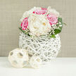 On a clean white surface, a lush, domed bouquet of white peonies and fuchsia-pink roses becomes the quiet focal point, illuminated by soft, balanced lighting that reveals every nuance of texture. The peonies sit at the centre, fully open and generous, their creamy ivory petals layered densely so they appear almost cloud-like, inviting thoughts of silk and gentle fragrance. Around them, the roses unfold in dramatic spirals of bright, saturated pink, their velvety petals adding depth and a passionate note to the composition. Between these larger blooms, sprigs of baby's breath form a delicate haze, tiny white flowers dotting the spaces and giving the arrangement a light, ethereal quality. Encircling the flowers, broad green leaves curve outward, polished and fresh, grounding the design with a natural, leafy frame. The bouquet is displayed in a unique round vase, crafted from interwoven white strands that mimic rattan or wicker, forming an open lattice that allows light to pass through and offers glimpses of stems within. In front of the vase, two pale woven spheres lie on the tabletop, one larger, one smaller, visually echoing the vase's airy, sculptural texture. Behind, a neutral beige backdrop with the look of finely textured fabric keeps the overall mood calm and refined, ensuring the vivid pinks and soft whites draw the eye. The scene feels like a snapshot from a Stanmore celebration, perhaps a bridal suite near Stanmore Country Park or a stylish home just off The Broadway, with flowers freshly delivered to mark a wedding, engagement, or heartfelt anniversary in quietly luxurious style.