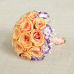 Captured from above and slightly to one side, this elegant bridal bouquet rests on a textured light beige fabric that resembles linen, the neutral ground sharpening each hue. A dense, rounded cluster of peach and soft-orange roses forms the bouquet's centre, their layered, velvety petals unfolding in perfect spirals that glow under soft, natural light. Scattered through the mass of roses are white lisianthus with frilled petals tipped in vivid violet, their two-tone edges lending a contemporary, artistic note that highlights the warmth of the roses. Delicate sprigs of baby's breath tuck into crevices between larger blooms, lending a whisper-light texture and a note of innocence to the overall silhouette. The stems are bound in a pale peach satin ribbon that catches the light and trails from the lower right, while a strand of cream pearls wraps gracefully around the handle, offering an heirloom touch. The fabric backdrop and the careful play of shadow create a sense of intimacy, as if the bouquet has been placed on a dressing table near Stanmore High Street while a bride prepares for her day. The scent of roses seems almost perceptible in the description-the soft, heady fragrance that accompanies such classic blooms. This arrangement reads as romantic and refined, a thoughtful choice for weddings and anniversaries that calls attention to careful colour pairing, airy texture, and the small, handcrafted details that make a floral piece feel personal and timeless.
