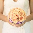 A close-up, chest-level photograph centres on a bride standing quietly, her torso framed against a soft, pale background that could be a light-filled Stanmore registry room or a calm corner of a local church. She wears a classic white or ivory wedding gown, the bodice adorned with delicate lace and a gentle V-neckline that hints at timeless bridal style. The fabric of the skirt falls in smooth, graceful folds beneath her hands, adding to the sense of poise and ceremony. Cradled carefully between her subtly manicured fingers is a round, hand-tied bridal bouquet that becomes the true focal point of the image. The bouquet is composed mainly of tightly clustered roses in shades of peach, light apricot, and blush pink, each bloom full and layered, the petals spiralling inward to create a soft, romantic dome. Beneath this cloud of warm-toned roses, a distinct ring of deep purple flowers edged with ruffled white-likely lisianthus-forms a contrasting halo, giving the bouquet depth and a slightly modern twist. Tiny white blossoms, resembling baby's breath, are woven delicately around the base, lending an airy, lace-like texture that echoes the gown. Her nails are painted in a light, neutral shade, with the faint shimmer of glitter on one thumb suggesting a personal, playful detail that might catch the light as she walks down a Stanmore aisle. The scene captures the quiet anticipation of a wedding moment: the bouquet held close to the heart, the colours soft yet confident, ideal for illustrating bridal floristry, wedding planning, or romantic colour palettes for ceremonies across Stanmore and neighbouring North London streets.