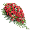 A dignified red funeral spray with a graceful, elongated form is shown against a bright white background, revealing each element with clarity. The spray is fuller at the upper left, where a dense cluster of blooms gathers, and then narrows in a soft, controlled cascade toward a delicate point at the lower right, forming a classic teardrop or casket-spray shape. The main flowers are deep red roses, their velvety petals fully open and arranged in layered spirals, conveying a sense of timeless love and quiet respect. Tucked among them are numerous red spray carnations, slightly brighter in tone, whose tightly packed, frilled petals create intricate texture and add fullness to the design. The vivid reds are framed by lush greenery: broad, dark green leaves anchor the arrangement and provide a strong outline, while soft, rounded eucalyptus leaves in a silvery green tone sit between blooms, easing the transition between flower and foliage. From the lower right corner and along the edge, delicate strands of lighter green trailing foliage extend outward, giving the impression of gentle movement and enhancing the cascading effect. The composition looks thoughtfully crafted, akin to a tribute prepared by Flower Delivery Stanmore for a service at a local chapel near Stanmore Station or along Marsh Lane. With its balanced blend of rich colour and natural greenery, this casket spray is well suited for resting on a coffin lid or standing at the front of a funeral or memorial, offering a heartfelt, visually comforting expression of sympathy, remembrance, and enduring affection from family and friends.