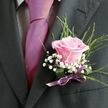 This image shows a refined wedding boutonniere on the lapel of a dark, impeccably cut suit, the centrepiece a single pale pink rose in tender bloom. The rose's petals are smooth and gently curved, tinged with a soft blush that grows slightly deeper toward the middle, suggesting freshness and care in its selection. Around the central flower, tiny white gypsophila blossoms create a soft halo, their delicate, cloudlike clusters adding dimension and contrast against both the rose and the sleek suit fabric. Slim strands of greenery extend outward in subtle arcs, bringing a natural energy to the arrangement that feels in tune with Stanmore's leafy avenues and nearby parkland. The stems are tightly wrapped in a narrow satin ribbon, a subtle lilac or mauve tone that harmonises with the rose's colour and adds a polished, formal finish. The lighting is calm and natural, illuminating the textures-the velvety petals, the fine gypsophila florets, the crisp weave of the lapel-without overpowering the scene. This boutonniere looks as though it has just been pinned moments before stepping into a Stanmore ceremony, perhaps with family gathering outside a local venue or synagogue. It is elegant without being ostentatious, relying on thoughtful composition and quality blooms rather than excess. You can sense the florist's trained eye in the way each element supports the others: the proportion between bloom and greenery, the choice of ribbon, the position on the jacket. It's a small but powerful accent, designed to sit close to the heart while promises are spoken and photographs capture the day's most cherished moments.