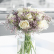 A calm, dignified bouquet of white roses and pale lavender filler flowers is captured here in a close-up view, evoking the kind of gentle sympathy arrangement often prepared for Stanmore families during tender moments. The roses are the main focus: generous blooms of creamy white, fully opened so that their intricately layered petals form soft spirals and rounded cups. Their surfaces appear velvety and smooth, with faint ivory undertones that lend warmth without disturbing the bouquet's soothing, neutral character. Nestled between the roses are slender stems of statice, carrying clusters of tiny, papery blossoms in shades of soft lilac, light mauve, and delicate pink. These small flowers add a mist-like, airy texture around the more substantial rose heads, creating a sense of quiet movement and depth without overwhelming the simplicity of the design. The arrangement is placed in a tall, slender clear glass vase filled with fresh, transparent water, through which the long green stems are clearly visible, standing in an orderly line that hints at the careful hand of an experienced florist. The vase sits on a smooth white surface that reflects some of the soft daylight, much like a table in a Stanmore home near Belmont Circle or along London Road. Behind the bouquet, the background fades into a gentle blur of whites and greys, leaving the flowers to speak for themselves. The overall feeling is one of respect, comfort, and understated beauty, suitable for offering condolences, marking a memorial, or simply bringing a sense of peace to a quiet corner in Stanmore.