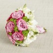 A vibrant bridal-style bouquet lies against a softly textured light beige fabric, captured from a slightly elevated angle so the rounded dome of flowers fills the frame. At the heart of the design are several large, fully opened roses in a romantic lavender-pink, their velvety petals deepening to a richer fuchsia tone at the outer edges, creating a gentle two-tone effect. Nestled between these roses are clusters of bright chartreuse green button chrysanthemums, their tiny, tightly packed petals forming crisp, spherical buttons that add playful texture and a fresh, contemporary pop of colour. Surrounding and softening the bolder tones are delicate white blooms, likely lisianthus, with layered, gently ruffled petals and subtle greenish-yellow centres that catch the light. A few closed green buds peek through, hinting at just-cut freshness, as if the bouquet has been gathered that morning from a Stanmore garden near Canons Park. The stems are neatly gathered and wrapped into a firm handle, finished with smooth pink satin ribbon that gleams softly in the diffused light. Over the ribbon, a fine strand of tiny white pearls spirals with careful precision, lending a touch of heirloom elegance reminiscent of wedding flowers carried in local Stanmore ceremonies. The overall mood is romantic yet modern, perfect for a bride walking through Bentley Priory surroundings, a special anniversary gift, or an intimate celebration at home, and reflects the detailed craftsmanship you might expect from a local florist offering hand-tied bouquet delivery across Stanmore and the nearby streets off Church Road.