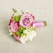 A small, hand-tied bouquet lies horizontally on a softly textured, light tan fabric, like linen laid across a Stanmore dining table before guests arrive. Two lush, fully open pink roses dominate the centre, their petals unfolding in tight, velvety layers that drift from gentle blush to deeper magenta tones at the outer edges, creating a natural ombré effect. Nestled between and around them are rounded tufts of lime-green button chrysanthemums, each globe made of countless tiny florets that bring playful texture and a vivid pop of colour. Creamy white filler flowers, reminiscent of lisianthus or petite freesias, weave through the design with their soft, layered petals, adding lightness and a sense of air moving through the bouquet. Tiny star-like white blooms with sunny yellow centres add further intricacy, like scattered confetti. The short stems are neatly gathered and bound with a smooth, fuchsia pink satin ribbon, its sheen catching the soft, even light. A fine strand of white faux pearls spirals delicately over the ribbon, evoking the quiet elegance of a wedding along Stanmore Hill or a celebration near Bentley Priory. The neatly trimmed green stems peek past the wrapping, reinforcing the freshly gathered feeling. The neutral background, with its visible weave, keeps the focus on the romance of pink, green, and white, making this compact posy feel perfectly suited for a bridesmaid, a young flower girl, or an intimate registry office wedding in Stanmore. It radiates poised, modern charm with a timeless, sentimental heart, as though it has just been set down for a moment between photographs and heartfelt toasts.