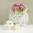 Captured against a softly blurred beige fabric backdrop, this floral centerpiece feels as though it has been placed on a freshly laid wedding table in Stanmore, perhaps in a hall near Canons Park. At the center sits a round, white woven vase crafted from slender, rattan-like branches that interlace to form an open globe, with gaps that keep the design light and airy. Rising from this sculptural vessel is a domed bouquet of roses and accent flowers. The roses are the stars: lush blooms in shades of pink and lavender, fully open so their petals swirl outward in generous, velvety layers. Many of the petals show a delicate deepening of pink at the edges, giving the arrangement a soft yet subtly dramatic look. Threaded between the roses are bright green button chrysanthemums, their tiny, tightly packed petals forming neat, spherical heads that punctuate the pastel tones and add lively texture. Around the lower edge, delicate white filler flowers slip between the larger blossoms, creating a gentle, cloud-like fringe that makes the bouquet appear abundant without feeling heavy. On the clean white surface beside the vase, two decorative spheres made from pale wicker or twigs echo the vase's woven texture, one larger and one smaller, adding elegance and balance to the composition. The neutral background, reminiscent of linen, keeps the attention firmly on the interplay of pink, lavender, green, and white. Overall, the image suggests refined romance and thoughtful craftsmanship, ideal as a wedding table arrangement, bridal luncheon centerpiece, or sophisticated home accent for celebrations across Stanmore.