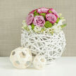 On a pristine white tabletop that offers a faint mirror of every curve and stem, a contemporary floral arrangement glows softly, as if ready to be carried into a Stanmore reception venue near Belmont Circle. The centerpiece rises from a unique spherical vase composed of slender, white-washed branches, woven together in an open, lattice-like pattern. This handcrafted vessel feels light and modern while echoing the natural textures of nearby Stanmore Country Park. Above it, the flowers form a rounded, harmonious dome. Lush pastel pink roses dominate the upper section, each bloom fully open, with velvety petals spiralling outward and some edged in a deeper rosy hue. Among the roses, clusters of bright lime-green button chrysanthemums tuck into the gaps, their small, rounded flower heads adding structure and a refreshing pop of colour. Circling the base, a ring of delicate white blossoms with tiny yellow centres spills just past the rim, creating a soft fringe that eases the transition into the woven vase below. To the left of the main arrangement, two decorative balls crafted from pale wooden strips lie casually on the table, one noticeably larger than the other, echoing the airy geometry of the vase and tying the elements together. The background is a warm, neutral taupe wall with gentle texture, blurred just enough to keep attention focused on the bouquet. Bathed in natural light, the petals appear dewy and fresh, suggesting flowers arranged and delivered the same day by a skilled local florist. The mood is serene yet celebratory, suited to weddings, engagements, or meaningful gatherings throughout Stanmore and nearby Harrow.