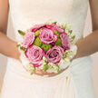 Held close against the intricate detail of a bridal gown, this bouquet feels like a graceful pause before the walk down the aisle in Stanmore. At the heart of the arrangement, lush lavender-pink roses unfurl confidently, their petals velvety and dense, arranged in gently rounded heads that seem made for close-up photographs on The Broadway or in a nearby garden. Their tone sits delicately between mauve and dusty blush, a flattering, romantic shade that pairs beautifully with ivory dresses. Tucked among these classic roses are zesty green button chrysanthemums, their compact, domed blooms adding an unexpected splash of contemporary energy. Their lime hue lifts the entire palette, ensuring the bouquet feels joyful and modern rather than overly traditional. Around the edges, clusters of creamy white flowers and slender buds form a soft, cloud-like halo, echoing the lightness of tulle veils and lace sleeves seen in Stanmore's bridal boutiques. The design is round and securely hand-tied, so it sits comfortably in the bride's hands, inviting her to hold it close during those quiet, anticipatory moments before the ceremony. Soft, natural light glides over each bloom, catching the subtle sheen of the rose petals and the textured surfaces of the chrysanthemums. The imagined scent is subtle yet comforting: the familiar sweetness of fresh roses interwoven with crisp green stems. Every element has been chosen with care and local expertise, reflecting a florist's understanding of which flowers will stay radiant from the first photos to the final farewell. This bouquet speaks of joy, hope, and new beginnings in Stanmore, captured in colour, texture, and gentle fragrance.