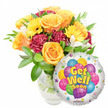 A bright, uplifting get-well gift captured against a clean white backdrop: a lush, hand-arranged bouquet sitting in a clear glass vase beside a shiny round Mylar balloon that reads Get Well Soon. The flowers are centered slightly left, with several large, softly unfurling roses in warm orange and deep yellow forming the visual heart. Golden-yellow gerbera daisies with sunlit faces push forward like small suns, while rich burgundy-to-maroon carnations add ruffled texture and shadowed depth between the rose blooms. Sprays of delicate white alstroemeria thread through the composition, their freckled throats and slender petals lending a light, airy contrast to the denser blooms. Glossy green foliage-broad leaves and finer stems-wraps the arrangement, framing each flower and giving the display a fresh, just-delivered look; the clear vase reveals the stems and water, hinting at the florist's careful conditioning. To the lower right, a metallic silver foil balloon partially overlaps the bouquet: its glossy surface reflects soft studio light, while a cheerful cluster of cartoon-style balloons in blue, purple, green, pink, and yellow is printed at the center, some patterned with stripes, polka dots and checkerboards. Bold yellow-and-blue lettering with a subtle shadow declares the message, and tiny starburst motifs dot the white border; at the very bottom edge a small KALEIDOSCOPE logo is visible. The overall impression is one of warmth, optimism and gentle care-an arrangement that smells faintly of citrus roses and green leaves, meant to comfort someone convalescing and to brighten a bedside or kitchen table in Stanmore or nearby Canons Park.