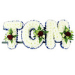 A personalised floral tribute spelling the name TOM is shown here in crisp focus, the three block letters arranged horizontally on a slim dark frame so they appear ready for display at a funeral or memorial service in Stanmore. Each letter is completely covered with tightly packed white chrysanthemums, creating a soft, cushion-like texture of overlapping petals that conveys purity and peace. Around the outside edge of every letter runs a royal blue satin ribbon, neatly folded and pinned to follow each straight line and curve. Its smooth, reflective surface offers a rich contrast that clearly defines the name against the plain white background, similar to the traditional tributes often seen at services around Stanmore and Harrow Weald. Carefully positioned at the upper sections of the 'T', the curve of the 'O', and the shoulder of the 'M' are elegant floral clusters that add depth and colour. Deep, velvety red roses sit at the heart of each cluster, surrounded by smaller white or cream spray roses and slender white blooms evocative of freesia, which introduce a delicate vertical rhythm and a gentle fragrance. Around these sit sprays of fresh green foliage-feathery ferns and textured leaves-that bring movement and a natural, garden-like feel, as if the arrangement carries a whisper of the greenery from Stanmore Country Park. The overall effect is one of quiet dignity and heartfelt remembrance, a bespoke floral name arrangement that honours Tom with love, making it a fitting choice for families in Stanmore seeking a meaningful, named funeral tribute.