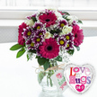 A charming combination of flowers and a heart-shaped balloon is shown here as though it has just been placed on a dining table in a Stanmore home, ready to delight its recipient. The focal point is a lush bouquet arranged in a rounded, clear glass vase, where straight green stems are visible beneath the surface of clean water, adding to the sense of freshness and care. Tied around the neck of the vase is a length of rustic natural twine, knotted in a simple bow and holding a small, rectangular white tag. The tag carries a delicate pink rose motif and green, hand-written-style wording, evoking the feel of a personal note attached to a lovingly prepared gift. Above the vase, the bouquet forms a full, rounded dome. Several large gerbera daisies in a rich magenta-pink hue command attention, their perfect circles of smooth petals framing deep, dark centres that lend drama. Surrounding them are clusters of smaller chrysanthemums, whose petals are a striking gradient from velvety purple at the base to clean white at the tips, arranged around bright yellow centres that glow warmly. Creamy white roses are nestled throughout, their soft, layered petals associated with romance and gratitude, while delicate sprays of white baby's breath lace through the arrangement, adding a light, airy touch. Fresh green foliage fills in the spaces, creating depth and a natural, garden-inspired look that feels at home in the greenery around Canons Park and Stanmore Hill. To the right of the vase, a shining heart-shaped foil balloon leans gently toward the flowers. It features a reflective silver edge and a white centre decorated with playful, multi-coloured letters in shades of pink, purple, and blue that spell out Love n' Hugs 24-7, with tiny hearts scattered among the wording. Resting on a smooth white surface with a softly blurred, bright background, this image captures the mood of a joyful flower delivery in Stanmore, perfect for birthdays, anniversaries, or sending ongoing love and hugs from morning to night.