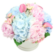 The scene shows a luxurious pastel floral hatbox arrangement, designed with the kind of quiet sophistication often appreciated in Stanmore's elegant homes and apartments near the Broadway. A round, immaculate white hatbox sits centrally against a white background, its subtle ridge detailing framing a lush cloud of blossoms that rises in a generous, mounded form. The arrangement feels rich yet serene, with no visible gaps between the flowers, as if every stem has been placed with intention. Large, soft pink peonies form the visual anchor, their multi-layered petals unfolding in delicate ruffles that give a sense of romance and abundance. Threaded between them are rounded heads of light blue hydrangeas, whose countless tiny florets create a pillowy texture and cool, soothing colour that balances the sweetness of the pinks. Scattered throughout these larger blooms are petite light pink spray roses, nestled closely so that buds and half-open blossoms add intricate detail and a sense of gentle unfolding. Two striking pink calla lilies rise gracefully from the pastel dome, one on the upper left and one to the right, their smooth, sculpted trumpets stretching slightly above the rest of the flowers and adding height and a contemporary touch. A few deep green leaves peek discreetly from the lower edges of the bouquet, grounding the composition and offering refreshing contrast. The lighting is soft and even, allowing each petal and curve to appear crisp yet delicate, like a calm morning in Stanmore before the day begins. The overall feeling is one of tender luxury and thoughtful care, perfect for welcoming a new baby, sending heartfelt congratulations, or offering gentle comfort to someone living near Stanmore station or along its quiet, tree-lined roads.