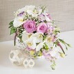 A romantic floral centrepiece sits in a rounded white wicker basket on a bright white surface, styled in a way that would perfectly suit a Stanmore wedding reception or anniversary dinner. The arrangement is rich with pink and white blooms, beginning with fully open pastel pink roses whose layered, spiralling petals create a soft, luxurious heart to the display. Nestled among them are graceful white Phalaenopsis orchids, each flower presenting wide, flat petals that curve gently back from vivid yellow-orange centres with subtle markings, lending a refined, modern elegance. Encircling these focal blooms are white lisianthus with delicately ruffled edges, some petals brushed with a faint hint of lavender-purple that adds a cool, understated contrast. Pale pink stock flowers rise on slender stems, their clustered blossoms providing height and a gentle sense of movement that recalls the breeze over Stanmore's green spaces. Fine threads of baby's breath weave between the larger flowers like a light mist, softening transitions and adding a delicate, cloud-like texture. Pockets of fresh green foliage, with smooth, glossy leaves, peek between blooms and around the base, grounding the pastel palette in nature and providing depth. The white wicker basket itself is tightly woven, its round shape and subtle texture offering a rustic yet refined base. On the tabletop beside it lie two decorative spheres formed from interlaced strips of pale natural rattan, echoing the basket's pattern and enhancing the organic, handcrafted feeling. Behind everything, a softly textured beige backdrop similar to linen provides a warm, neutral canvas that lets the colours and shapes of the flowers stand out. The arrangement radiates calm joy and intimate celebration, ideal for marking special moments across Stanmore.
