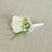 A single white orchid boutonnière rests on a pale, linen-like fabric, presented as though ready to be pinned to a sharply tailored suit before a ceremony in Stanmore. The orchid, luminous and unblemished, opens in broad, velvety petals that curve gently outwards, their surface smooth and almost reflective in the soft, even light. At the center, a subtle glow of buttery cream and pale yellow draws the eye inward, giving depth and warmth to the otherwise cool, snowy bloom. Surrounding this focal flower is a light wreath of baby's breath, each tiny white blossom hovering on fine stems like a mist of delicate dots, echoing the orchid's purity but on a smaller, more intricate scale. Slender strips of vibrant green foliage slip out from beneath the main flower, their narrow blades suggesting movement, as if freshly cut from a garden near Canons Park or one of Stanmore's leafy back gardens, bringing a refreshing contrast to the white on white. The stems are carefully bound together and enveloped in a gleaming white satin ribbon, wrapped smoothly and tightly so there are no loose ends, only a polished, cylindrical handle that feels comfortable to hold and easy to secure. A petite satin bow sits near the top of the binding, adding a hint of formality and craftsmanship, like the finishing touch a local florist at Flower Delivery Stanmore would give just before sending the piece out for same-day wedding delivery. The backdrop fabric, a warm light tan with a fine woven texture, lends a calm, understated foundation, allowing the boutonnière's elegance, soft fragrance, and air of quiet celebration to take center stage.