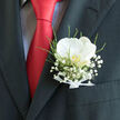 In this close-up image, the focus rests on the pocket of a dark charcoal suit, where a pocket boutonnière has been carefully placed to mark a day of celebration in Stanmore. The central bloom is a striking white orchid, its petals thick, smooth, and gently curved, giving it a sculptural elegance that stands out against the dark fabric. The creamy tone of the orchid glows softly, with a discreet wash of pale gold in the centre that hints at warmth and intimacy, like the glow of late afternoon light over Stanmore Common. Encircling the orchid, tiny clusters of baby's breath float in a delicate halo, each minute flower lending an almost ethereal softness, as if the blossom rests within a cloud. Slender green sprigs slip between the white flowers, adding texture and a whisper of movement, reminiscent of the quiet rustle of leaves along the streets leading to Stanmore station. At the base of the design, a slim white ribbon is tied around the stems with almost ceremonial care, its satin sheen catching the light in smooth, subtle highlights. Beside the boutonnière, a bold red tie introduces a vivid contrast, its glossy silk amplifying the orchid's freshness and emphasizing the formality of the occasion. The interplay of soft blooms and structured tailoring, bright white and deep charcoal, conveys a sense of balance between romance and poise. This boutonnière feels like a bespoke choice for a wedding, engagement, or milestone event in Stanmore, symbolizing love, elegance, and the quiet emotion of a meaningful moment.