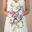 A bride stands elegantly, framed from mid-chest down, the warm softness of a pale tan background lending a gentle serenity reminiscent of a summer afternoon in Stanmore. She gently holds an extraordinary cascading bridal bouquet, the heart of the image and a showpiece of floral design as found at local florists in the area. The bouquet descends in a graceful teardrop cascade, dominated by large, pristine white phalaenopsis orchids whose broad petals and buttery centers evoke refined luxury and quiet splendor. Interwoven are blooming blush-pink garden roses, unfolding their romantic layers between clusters of lavender stock and delicate lisianthus, some rimmed in irresistible violet edging. Wispy sprays of white baby's breath add airy fullness, letting the bouquet breathe, while just a trace of greenery nods to Stanmore's tree-lined streets. The bride's arms, gently curving around the elegant arrangement and her strapless, lace-embellished gown, serve as natural frames for the flowers' artistry. Every petal and flourish whispers of joyful anticipation and bridal elegance. This bridal bouquet is an inspired reflection of the distinctive style and beauty cherished in Stanmore weddings-an arrangement that feels as timeless and luminous as the community itself.
