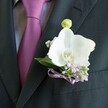 A close-up photograph captures a finely tailored charcoal suit jacket worn for a special occasion in Stanmore, with the camera lingering on the left lapel where an elegant white orchid boutonniere is pinned. The fabric of the jacket is smooth and dark, its weave subtly visible in the soft, diffused light, while a mauve-magenta tie peeks out from beneath the lapel, neatly knotted over a pale grey dress shirt with a delicate texture. At the heart of the image is a pristine Phalaenopsis orchid, its broad, satiny white petals curving outward like sculpted silk, framing a soft yellow and warm orange centre that draws the eye. Around the orchid, you can see tiny sprays of baby's breath and clusters of pale lilac blossoms, perhaps heather or a similar airy flower, which add a gentle haze of colour and texture, reminiscent of spring borders in Stanmore Country Park. A slender lavender ribbon is wrapped carefully around the stems, tied into a neat bow that rests just above the suit's breast pocket, echoing the tones of the tie and creating a sense of deliberate coordination. Above the main bloom, a small green orchid bud stands poised to open, hinting at new beginnings and quiet anticipation. The arrangement feels fresh and thoughtfully composed, like something a local florist would create for a groom stepping out onto Church Road or heading to a wedding at a nearby Stanmore venue. Every detail-from the sheen of the petals to the soft contrast of dark cloth and light flowers-suggests understated romance, calm confidence, and the intimate beauty of a moment that will be remembered long after the petals have faded.