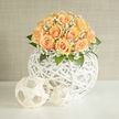 A masterfully balanced bouquet of peach-honey roses and white gypsophila sits poised on a modern white woven orb, radiating quiet celebration and refined craftsmanship. The roses, blushed with soft apricot tones, form a domed cluster whose layered petals fold inward in delicate spirals, each bloom reflecting a hint of golden light across its satiny surface. Interwoven tufts of baby's breath create a soft, cloud-like border around the roses, diffusing the composition and lending a bridal, ethereal softness that contrasts beautifully with the crisp geometry of the rattan sphere. Green foliage peeks through in measured amounts, offering cool colour contrast and a living freshness that balances the overall warmth. The white wicker vessel-light, architectural, and open-lets shadow play across the tabletop, producing a quiet rhythm of lines that complements the blooms' organic curves. Imagine the faint scent of fresh petals and powdered florals, a scent that suggests tenderness and new beginnings, perfect for a wedding table, a heartfelt congratulations, or a calm birthday gathering. As a local arrangement often requested around Stanmore High Street and Canons Park, this piece suits neighbourhood celebrations and small receptions alike. Flower Delivery Stanmore prepares similar arrangements with attention to balance and longevity, choosing stems at peak bloom so the bouquet maintains its soft luminosity for days. Overall, the design reads as contemporary yet timeless-an elegantly understated statement that welcomes both the eye and the hand.