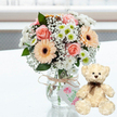 A warm and affectionate gift set is arranged on a smooth white surface, evoking the feeling of a carefully prepared anniversary surprise in Stanmore. At the centre stands a clear glass jar-style vase filled with a rounded bouquet of fresh flowers. Blushing pink roses open gracefully alongside peach-toned gerbera daisies, whose open faces are accented by distinct, dark brown centres. Around them, light sprays of white baby's breath blur into a soft haze, while neat white chrysanthemums with bright lime-green centres add structure and a fresh, garden-like vibrancy. The stems are visible through the glass, and a length of natural twine wraps around the neck of the vase, tied in a simple bow. From this bow hangs a small, rectangular white tag featuring a pink heart symbol and the word Handmade in flowing green script, hinting that this bouquet has been composed with individual attention, like the custom creations often sent around Stanmore's quiet residential streets and parks. To the right of the vase, a cream teddy bear sits upright on the same faintly reflective tabletop. Its plush fur appears soft and slightly textured, with dark bead eyes that catch the light, a tiny brown nose, and a subtle stitched smile. A narrow dark brown satin ribbon is tied neatly around its neck, echoing the tones in the gerbera centres. The background is out of focus and bright, suggesting daylight filtering through a nearby window, perhaps in a Stanmore home overlooking local greenery. The overall impression is one of tenderness, stability, and gentle celebration, ideal for marking anniversaries, sending comforting wishes, or simply showing you care within the Stanmore community.