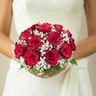 In this detailed bridal close-up, a hand-tied bouquet of luxurious red roses and airy white baby's breath rests against a smooth white wedding dress, evoking the refined yet heartfelt celebrations often seen in Stanmore's churches and halls. Each rose is a deep, velvety crimson, its petals layered in a perfect spiral that draws the eye inward toward the centre, like a little whirlpool of colour and emotion. The blooms are clustered snugly into a rounded, almost sculpted shape, creating a sense of fullness and intention-as if every flower was chosen with care after a thoughtful chat with a local florist near Stanmore station. Surrounding the roses, clouds of gypsophila weave in and out, their countless tiny white florets softening the bouquet's outline and lending it a delicate, romantic haze. The contrast is striking yet soothing: vivid red and crisp white, with flashes of fresh green foliage nestled between stems, adding a touch of natural freshness that grounds the design. The lighting is gentle and flattering, highlighting the texture of each rose petal and the fine, frothy detail of the baby's breath, much like the soft daylight that spills into a reception room at The Hive or a nearby country house venue. Even without scent, the image suggests fragrance-the familiar, enveloping perfume of red roses mixed with the subtle greenery notes that accompany freshly cut stems. This bouquet feels timeless and deeply personal, the kind of arrangement a bride might carry proudly down an aisle in Stanmore, knowing that its classic beauty and rich symbolism of love, purity, and devotion will translate effortlessly into every photograph and cherished memory.