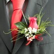 This close-up image captures an elegant wedding buttonhole pinned carefully to the lapel of a dark charcoal grey suit jacket, evoking a groom or groomsman about to step into a Stanmore ceremony. Central to the arrangement is a partially opened rosebud in a vivid fuchsia pink, its tightly wound petals forming a refined, velvety swirl that draws the eye. Around the rose, delicate clusters of white gypsophila-baby's breath-create a soft, cloudlike outline, each tiny flower standing out clearly against the deeper background of the suit. Fine, feathery strands of greenery mingle with slightly broader, glossy green leaves, giving the boutonniere lightness and subtle movement, as though inspired by the calm greenery of Stanmore's residential streets and nearby parks. At the base, a small, bright red satin bow is tied with precision, its sheen and colour echoing the smooth, unpatterned red silk tie that peeks from beneath the lapel. The tie lies against a subtly patterned grey dress shirt, and the close-woven wool blend of the suit suggests quality and formality. Gentle, even lighting allows every texture to be seen clearly, from the softness of the petals to the crispness of the foliage and the lustre of the ribbon. The overall effect is both romantic and composed, a carefully coordinated buttonhole suited to weddings, anniversaries, or special events around Stanmore, Edgware, and the surrounding area, where thoughtful floral details help to complete a polished, memorable look.