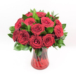 A luxurious bouquet of deep crimson red roses fills the frame, each bloom densely clustered to create a rounded, opulent arrangement that feels almost like a living jewel. Every rose head is perfectly formed, with velvety petals spiralling from a tight, secretive centre into wider, softly unfurling edges that catch the light. The red is intense and ruby-like, a saturated tone that suggests passion, devotion, and classic romance, much like the timeless gestures often exchanged around Stanmore Hill or along The Broadway on special evenings. Threaded between the blossoms is a generous collar of rich, dark green foliage, likely Ruscus or a similar glossy leaf, which frames the roses and sharpens their colour with its cool, fresh contrast. The leaves appear smooth and slightly waxy, some catching a gentle sheen that adds depth and movement to the composition. All of this beauty rises from a translucent ruby-red glass vase, its surface etched with a graceful twisted or spiralled pattern. The vase narrows at the waist and flares slightly at the rim, giving the arrangement a poised, elegant stance, as if ready to be placed on a dining table in a Stanmore townhouse for an anniversary dinner. Through the coloured glass, the straight rose stems can be seen, layering shades of red and green that echo the bouquet above. Set against a pristine white background and illuminated by bright, even lighting, every petal, leaf, and curve is crisp and vivid. The result is an image that radiates romance and sophistication, perfectly suited to moments of love, gratitude, or heartfelt celebration shared quietly at home or after a walk in nearby Canons Park.