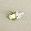 A classic white rose wedding boutonniere lies diagonally across a softly textured, light beige fabric that resembles natural linen, its fine crosshatch weave giving the scene a calm, understated elegance often favoured for Stanmore ceremonies. At the heart of the design is a single creamy white rosebud, its velvety petals still tightly furled yet gently opening, suggesting a fresh bloom chosen early that morning. Around the rose, tiny clusters of white baby's breath form a delicate halo, their cloud-like blossoms creating a soft, airy contrast against the smooth rose petals. Slender, pointed deep green leaves and a few feathery, fern-like sprigs cradle the central flower, adding depth and a quiet woodland freshness that feels at home in the leafy streets near Stanmore Country Park. The stems are neatly gathered and wrapped in a pristine white satin ribbon, the texture smooth and gently reflective in the diffused light. Just beneath the bloom, the ribbon is tied into a small, tidy bow, indicating that the piece is ready to be pinned to a groom's lapel for a church wedding on Marsh Lane or an elegant reception near Canons Park. Soft, even lighting falls across the arrangement, emphasising every fold of the rose, each tiny speck of the gypsophila, and the clean lines of the foliage, while the neutral background keeps all attention on the floral craftsmanship. The image conveys quiet anticipation and polished sophistication, the kind of timeless boutonniere a local florist in Stanmore might prepare for groomsmen, pageboys, or a formal black-tie event, capturing the symbolism of purity, new beginnings, and refined, classic style in a single, carefully composed detail.