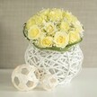 A sophisticated floral display sits calmly on a pure white tabletop, evoking the mood of an intimate celebration in Stanmore. At its centre rises a carefully rounded bouquet of pale yellow roses, each flower fully open yet still holding its cupped, sculptural form. Their petals overlap in soft layers, giving the impression of a velvety, sunlit cushion. Between these roses, fine sprays of white gypsophila weave through the arrangement, their clusters of tiny blossoms adding lightness and a dusting of cloud-like texture that softens the dome. Around the base of the flowers, an elegant, slender band of rich green leaves encircles the stems, offering a touch of contrast and highlighting the boundary between blooms and vase. The arrangement is housed in a distinctive white woven vase with an open, basket-inspired design. Thin, curved strands interlace to form its body, allowing light to pass through and cast delicate patterns onto the glossy white surface below. Resting beside the vase are two small decorative spheres in a pale, natural wood tone, each fashioned from intertwined strips that mirror the vase's airy latticework and add a sculptural note. Behind the arrangement, the background is softly blurred in a warm beige tone, reminiscent of a serene interior wall in one of Stanmore's quiet, well-kept homes, ensuring all attention rests on the flowers and their textures. The overall effect is one of gentle luxury and quiet joy, an ideal centerpiece for wedding tables, milestone celebrations, or as a tasteful floral gift delivered to loved ones across Stanmore by Flower Delivery Stanmore.