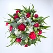 A richly detailed, top-down photograph shows a generous hand-tied bouquet spread like a colourful circle against a pure white background, making every bloom stand out clearly. At the centre, velvety deep red roses, some still in tight bud and others just beginning to unfurl, create a sense of romance and quiet luxury, their petals layered like satin folds. Around them nestle ruffled carnations in vivid fuchsia and ruby red tones, their frilly edges adding movement and a slightly playful, celebratory feel, as if the flowers are mid-dance. Clusters of dainty white spray chrysanthemums, each bloom with a sunny, daisy-like face, punctuate the richer hues, bringing brightness and a fresh, almost crisp calmness to the composition. Floating between these larger heads is a fine haze of tiny white gypsophila, or baby's breath, its countless miniature florets giving the illusion of a soft floral mist. The flowers are cradled by a lush frame of greenery: strong, pointed dark green leaves, likely from lily or alstroemeria stems, provide a bold architectural base, while rounded, silvery-green eucalyptus leaves weave through with a cool, contemporary touch and an imagined gentle, herbal scent. The bouquet appears full, balanced and expertly composed, the shapes and tones carefully layered so no flower feels crowded. It radiates warmth and affection, perfectly suited to send love, celebrate a milestone, or lift someone's spirits on a grey Stanmore afternoon, whether it's arriving at a home off the Uxbridge Road or a small office near Stanmore station, bringing colour, fragrance, and a heartfelt message without a single word spoken.