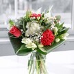A contemporary glass-vase arrangement brimming with emotion, this bouquet presents a striking duet of a coral-red rose and a plush crimson carnation, their velvety petals layered and full, almost glowing against a backdrop of snowy white blooms. Clusters of delicate white chrysanthemums mingle with daisy-like flowers and airy sprays of baby's breath, giving the design a soft, clouded halo that feels both lively and soothing. Between these, creamy white lily buds rise on tall, elegant stems, still closed and sculptural, suggesting new beginnings and beauty yet to unfold. The base of the bouquet is cradled by broad, deep-emerald leaves, their glossy surfaces carefully folded and curved to create a sleek, modern collar that frames the flowers and adds a sense of structure. Light streams in from a nearby window, the kind of gentle brightness you might find in a quiet Stanmore sitting room, catching the glass vase and casting subtle reflections on the table beneath. The overall mood is one of thoughtful celebration and calm joy, as if the arrangement has been prepared for a special birthday brunch near Stanmore Hill or a heartfelt thank-you delivered to a home off The Broadway. You can almost sense the fresh, green fragrance of cut stems and the faint sweetness of the rose and lilies, a clean, uplifting scent that suits both everyday moments and meaningful occasions. Crafted with care, this bouquet feels like a personal gesture of warmth, designed to brighten a space without overwhelming it, and to bring a gentle, enduring smile to whoever receives it in and around Stanmore.
