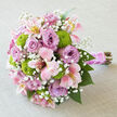 Placed horizontally on a pale grey, linen-textured cloth, this bouquet looks poised and serene, as though waiting to be lifted by a bride stepping out in Stanmore. The blooms are gathered closely into a rounded, compact shape, giving the arrangement an opulent, full-bodied appearance. At the heart of the display are numerous lavender roses, many fully open, their silky petals curling in layered spirals that invite the fingertips and suggest both refinement and heartfelt emotion. Threading between them, pink and ivory alstroemeria add movement and delicate interest, their slim petals enhanced with soft washes of yellow and fine, darker streaks that resemble hand-painted brushstrokes. Dotted around the bouquet like little jewels are bright chartreuse green button chrysanthemums, their perfect spherical forms and crisp texture giving a contemporary lift and a refreshing burst of colour. Wispy stems of white baby's breath weave throughout, their tiny blossoms forming a misty veil that softens the bold forms and adds an airy, romantic quality, similar to the gentle haze over Bentley Priory Nature Reserve on a sunny afternoon. The stems are expertly gathered and wrapped in a wide band of smooth pink satin ribbon, which encases the handle in a glossy, polished embrace. Over this satin, a delicate string of small white pearls is wound, giving the bouquet a sophisticated, bridal finish, like an heirloom bracelet. Below the binding, the natural stems extend in a straight, tidy line, offering a rustic balance to the luxurious blooms above. The overall impression is one of calm joy and thoughtful artistry, ideal for weddings, engagement celebrations or special occasions in and around Stanmore.