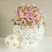 Infused with the understated grace typical of Stanmore, this exquisite floral centerpiece rests on a luminous white tabletop, softly illuminated as if by mid-morning sun filtering through leafy garden windows. The arrangement is anchored by a round, openwork vase-its white woven lattice recalling the craftsmanship of local artisans, sturdy and beautiful in equal measure. Inside, pastel-pink and lavender roses blossom in full, velvety layers, their romance heightened by the presence of delicate trumpet-shaped pink alstroemeria, streaked with subtle inner markings. Bright, spherical lime-green button chrysanthemums add a spirited, playful twist, nestled amongst light drifts of baby's breath which imbue the bouquet with an airy elegance. Subtle swathes of dark greenery peep between blooms, their richness reminiscent of Stanmore's landscaped gardens. To the left of the arrangement, two decorative spheres in pale natural tones-one large, one small-sit gracefully side by side, their woven forms echoing both the rustic chic of the vase and the traditions of Stanmore's artisan markets. Against a soft beige background with gentle texture, every element invites pause and admiration. Perfect as a wedding centerpiece, table adornment, or artistic accent, this display evokes freshness, optimism, and the timeless beauty that defines special moments in Stanmore.