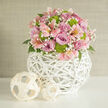 A serene and sophisticated floral scene unfolds in this close-up, reminiscent of a carefully arranged wedding display in a Stanmore home or boutique venue along The Broadway. At the heart of the composition is a beautifully balanced, rounded bouquet resting in a distinctive white woven vase shaped like a small globe. The vase's delicate wicker strands intersect to form a light, airy lattice, giving the base a contemporary, sculptural presence. Rising from this modern pedestal, an abundant cluster of roses in shades of lavender and soft rose pink creates a plush, romantic centre. Their petals gently curl outward, revealing spirals that feel almost like soft fabric, and a few just-opening buds add a sense of anticipation and new beginnings. Interspersed throughout are graceful stems of pale pink alstroemeria, their slim petals traced with darker speckles and fine stripes, like tiny brushstrokes that catch the eye without overwhelming the design. Vivid lime green button chrysanthemums nestle between the pastels, their neat, domed shapes giving rhythm and a fresh, energising lift to the palette. Threading delicately through every layer, dainty clusters of white baby's breath introduce a cloud-like softness, blurring the boundaries between one bloom and the next. Peeks of dark green leaves add depth and ensure the bouquet feels lush and garden-inspired. The arrangement sits atop a pristine, smooth white surface that reflects a gentle glow of ambient light. To one side, two smaller decorative spheres made from pale, flat woven strips mirror the vase's texture and form, tying the visual elements together. Behind everything, a neutral, lightly textured wall in beige tones keeps the background calm and unobtrusive. The overall impression is of quiet celebration and tasteful design, perfectly aligned with wedding collections or elegant gifts available for delivery across Stanmore.