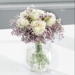 A close-up photograph shows a graceful bouquet of creamy white roses rising from a misty cloud of soft lavender statice, arranged in a clear, crackle-textured glass vase that glitters gently in the light. Each rose is in perfect condition, its velvety petals unfolding in spirals from a faintly buttery centre to a pure, milky edge, giving the blooms a fresh, just-opened look. Around them, countless tiny star-shaped flowers of pale purple statice create a feathery halo, their papery texture adding depth and a slightly rustic charm to the refined roses. The stems, a healthy, vibrant green, can be seen clearly through the water in the vase, criss-crossing like a subtle pattern and anchoring the design with a sense of natural structure. The vase itself has an ice-effect surface, like frozen water or cracked glass, catching stray glimmers of daylight and casting soft reflections onto the white tabletop beneath. Gentle shadows fall at the base, giving the arrangement a calm, grounded presence. The background fades into a blurred, pale grey, free from distractions so that all attention rests on the flowers and their interplay of white, green, and lavender. The overall mood is tranquil and soothing, like a quiet morning in a Stanmore sitting room looking out towards Bentley Priory Nature Reserve. This is the kind of arrangement you might have delivered to a friend recovering at home, placed on a bedside table, or used as a subtle, elegant centrepiece for a small family gathering, radiating peaceful beauty and understated care.