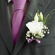 A sharply focused close-up of a groom's charcoal grey suit jacket captures the quiet luxury of formalwear on a wedding morning in Stanmore. The fine wool fabric has a subtle, almost herringbone texture that softens in the light, with the right lapel crisply pressed and perfectly aligned. At the heart of the image is a single, fully opened white lisianthus boutonnière pinned to the left lapel, its many ruffled petals layered like soft folds of silk. Two slender ivory buds rise just above the main bloom, still tightly closed, suggesting the promise of moments yet to unfold. Around them, a cloud of tiny white baby's breath stars outwards, each flower no bigger than a pinhead, creating a delicate halo against the dark jacket. Thin, airy strands of greenery arc gracefully behind the flowers, adding a sense of movement and freshness, like a whisper of breeze drifting across Canons Park. At the base, a narrow mauve ribbon is wrapped neatly around the stems and tied into a small, precise bow, its gentle sheen echoing the rich magenta tie that lies between the suit lapels. The tie itself is silk-smooth and confidently knotted, drawing the eye with its vivid colour against the patterned white and grey shirt beneath. The overall feel is one of calm anticipation and refined romance, as though the wearer is moments away from stepping into a ceremony at a Stanmore church or local venue. Every detail of the arrangement speaks of careful florist craftsmanship, considered styling, and the quiet joy of a special occasion.