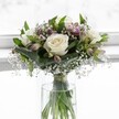 An elegant white rose bouquet stands in a clear cylindrical glass vase, bathed in soft, luminous light as if placed on a windowsill in a quiet Stanmore home. Three creamy white roses, fully open and velvety, form the calm heart of the arrangement, their petals unfolding in gentle spirals towards warm ivory centres that suggest freshness and purity. Around them, sprigs of gypsophila, or baby's breath, float like tiny white clouds, their miniature blooms scattered delicately through the design, creating a misty, romantic veil. Slim stems of alstroemeria in muted mauve and pale blush pink peek out between the roses and greenery, some still in bud and others just beginning to open, their softly streaked petals hinting at a tender touch of colour without disturbing the bouquet's overall serenity. Broad, rich green leaves are carefully folded and curved to cradle the flowers, adding structure and a deep emerald contrast that frames the pale tones beautifully. The stems are visible through the crystal-clear water of the vase, neatly gathered and tied just above the waterline with a subtle ribbon, a detail that speaks of thoughtful craftsmanship by a skilled local florist. The softly blurred white background heightens the sense of tranquillity, allowing the textures of petal, leaf, and glass to take centre stage. The bouquet feels perfectly suited to a peaceful moment of reflection, perhaps resting on a side table in a Stanmore living room after being delivered as a message of comfort, appreciation, or pure, uncomplicated love, its gentle fragrance and calm palette offering quiet reassurance.