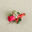 A richly detailed boutonniere lies on its side against a softly textured, light beige linen-style fabric, the kind of neutral backdrop you might see on a wedding morning in a Stanmore townhouse near the Broadway. At the heart of this small floral piece is a single, deep fuchsia pink rosebud, its velvety petals still tightly furled in the centre and gently loosening toward the edges, suggesting perfect freshness. Around the base of the rose, delicate sprays of white baby's breath cluster like tiny clouds, their minute blooms adding a lace-like softness and subtle contrast against the bold pink. Tucked between these white blossoms are glossy red hypericum berries, rounded and bright, bringing a cheerful, almost festive spark of colour that hints at celebration. The greenery is thoughtfully layered: fine, feathery, fern-like fronds extend in wisps from the sides, while broader, darker green leaves are curled and shaped to cradle the rose, giving the design structure and a sense of movement. The stems are carefully bound and hidden beneath a smooth wrap of rich red satin ribbon, tied into a small, neat bow that catches the light with a gentle sheen, echoing the romance of evening receptions at nearby venues around Stanmore Hill. Soft, even lighting falls from above, highlighting each texture-the matte linen, the satiny petals, the glossy berries, and the silkiness of the ribbon-so the craftsmanship of the boutonniere feels both intimate and refined, ideal for a groom, groomsman, or prom guest wanting a classic yet vibrant floral accent on their lapel.