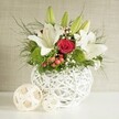 A romantic and refined floral centerpiece is presented as though ready to grace a wedding table in Stanmore, perhaps at a reception close to the green expanses of Bentley Priory. At the heart of the scene is a distinctive, spherical white vase made from smooth interwoven strands that create an open, basket-like pattern. It supports a carefully structured bouquet with a harmonious colour palette of white, red, and fresh green. Elegant white lilies tower above the arrangement, some already open with petals splayed wide in trumpet shapes, revealing delicate speckling and golden stamens, while others remain as elongated green buds, adding height and a sense of unfolding anticipation. Tucked among them, a single, flawless red rose becomes the emotional focal point; its velvety petals form a neat spiral, richly coloured and quietly dramatic against the surrounding white. At the base and nestled between taller stems, bright green pom-pom flowers-likely green dianthus or button chrysanthemums-provide mounds of soft, textured colour that feel joyful and contemporary, like the first new leaves in spring around Stanmore's residential streets. Fine clusters of white baby's breath thread between the main blooms, imparting an airy, cloud-like quality. Small red or reddish-brown berries punctuate the design with hints of festive warmth, making the centerpiece well suited to winter weddings or Christmas gatherings. A selection of greenery, from slender, feathery fronds to broader, darker leaves, fills out the silhouette and spills slightly over the rim, lending a natural, slightly untamed elegance. The vase rests on a bright white surface that reflects a gentle glow, and to its left lie two decorative spheres in pale, natural woven material, echoing the vase's lattice texture and completing the balanced composition. The background is a soft, neutral beige with subtle texture, allowing the lilies, rose, and greenery to stand out with calm sophistication for any special occasion in Stanmore.