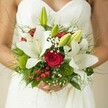 Delicately gathered and radiating gentle charm, this Stanmore bridal bouquet is a celebration of both romance and nature's artistry. Large, creamy-white lilies open widely within the lush composition, their petals smooth and luminous against the bride's textured white dress-echoing the fresh calm of Stanmore mornings. Interspersed between these blossoms are vibrant crimson roses, their velvety swirls drawing the gaze and evoking a timeless promise of love. Berries in vivid red add a playful and festive touch, while gossamer sprays of baby's breath drift between petals, softening the overall silhouette and lending a air of lightness and wonder. Mossy green orbs and graceful fern-like fronds complete the base, grounding the bouquet in a hand-gathered, natural style evocative of Stanmore's leafy landscapes. The bride's hands, visible at the bouquet's base, clasp the stems with quiet anticipation, as if drawing strength from the intricate textures and harmonious hues. Crafted to honour both joyful tradition and individual style, this bouquet stands as a tangible expression of new beginnings, perfect for local brides embarking on their wedding journey in Stanmore.