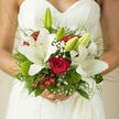 The photograph captures a hand-tied bridal bouquet nestled against a white lace wedding dress, its floral elements forming a rich tapestry of colour and texture that suits a Stanmore ceremony perfectly. At the heart of the bouquet, a single, full red rose draws the eye, its tightly layered petals rich and velvety, symbolising deep affection. Surrounding it, creamy white lilies bloom with poise, their broad petals opening in generous star shapes, some fully unfurled and others still in bud, hinting at stories yet to unfold like new journeys beginning near Stanmore station. Light clusters of gypsophila weave between the larger flowers, lending an airy softness, while bright red berries peek through the foliage, adding cheerful, jewel-like accents that echo the energy of wedding receptions around Marsh Lane or Uxbridge Road. Soft green moss and rounded dianthus add lush, cushiony texture at the base, grounding the arrangement with hints of nature, as though gathered from the edges of Stanmore Common. Surrounding foliage in multiple shades-from pale leaf-green to deep emerald-frames the central blooms and provides a verdant backdrop. Fine, trailing grasses arc gently above, creating an almost ethereal sense of movement as if stirred by a breeze during outdoor photos near Canons Park. The lace of the gown, visible behind the flowers, introduces delicate patterns that contrast with the smooth petals and glossy berries. Altogether, the bouquet appears thoughtfully composed, balancing tradition and a touch of the unexpected, embodying the quiet joy and graceful anticipation of a Stanmore bride on her wedding day.