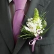 A close-up, high-angle photograph shows the upper torso of a man dressed for a formal occasion somewhere in Stanmore, perhaps just outside a wedding venue near Stanmore Hill. He wears a finely tailored charcoal grey suit jacket with a sharp lapel and clean shoulder line, set against a softly blurred background so the eye rests on his chest. Under the open lapels sits a pale dress shirt with a subtle grid pattern, perfectly pressed, and a smooth lavender necktie that falls straight down the centre, its sheen catching the light like satin under gentle indoor lighting. Pinned precisely to the left lapel is a handcrafted wedding buttonhole: a slim, pale green spiky flower bud rising upright, surrounded by open white alstroemeria blooms, each petal marked with delicate purple and maroon streaks and tiny golden stamens at their heart. Wisps of white gypsophila, or baby's breath, float between the lilies like a light cloud, while slender grass-like blades curl around the edges and broader deep green leaves cradle the blossoms. At the base, a neat lavender ribbon bow, perfectly colour-matched to the tie, secures the stems with a soft, satiny knot. The arrangement feels fresh, as though just prepared in a Stanmore florist's studio for same-day delivery, and the overall mood is one of restrained elegance and modern romance, capturing the quiet excitement of a groom or groomsman fastening his buttonhole before walking down the aisle. This detailed image celebrates the artistry of buttonhole design, from texture and tone to the careful harmony of white, green, and lavender.